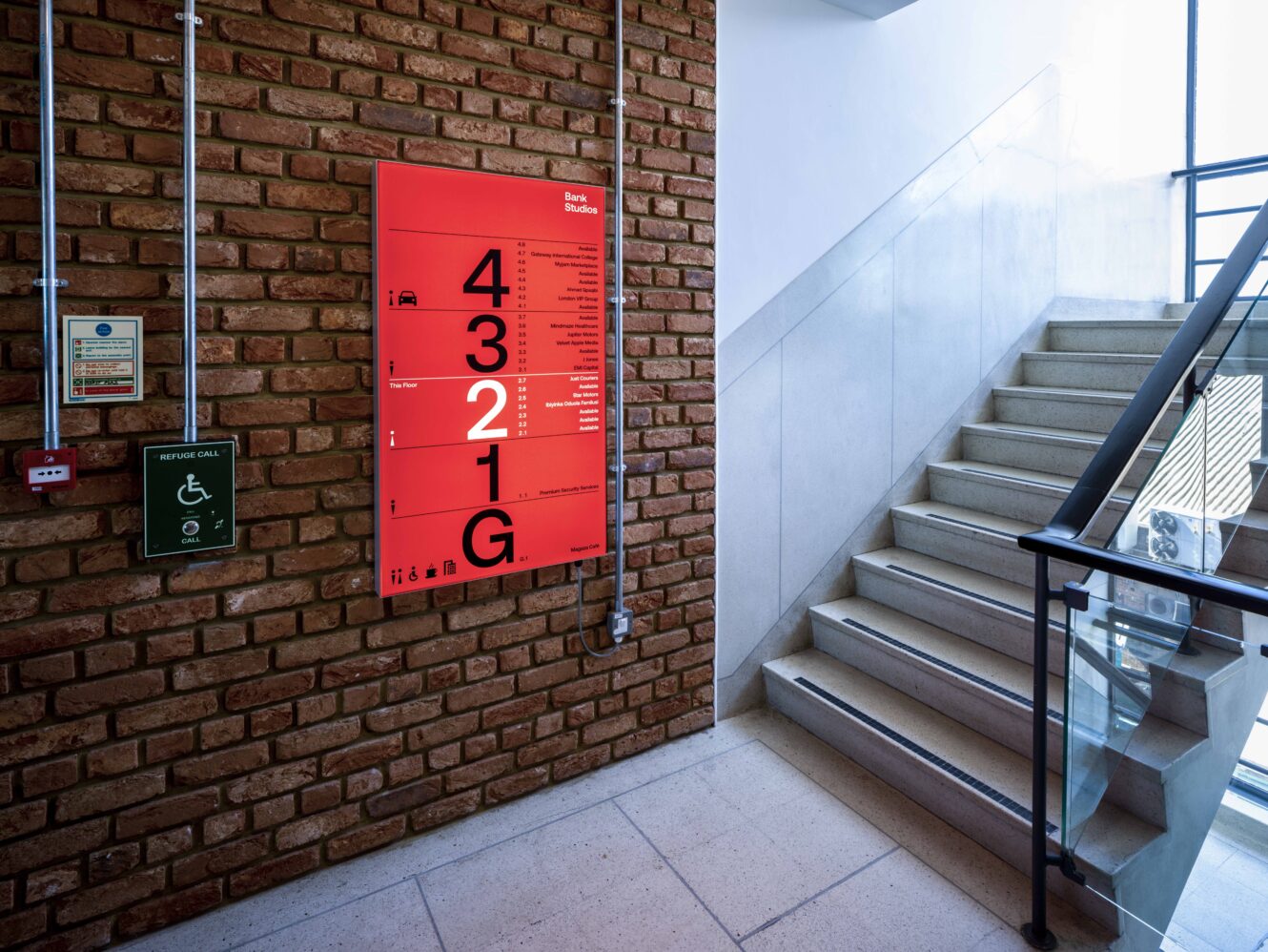 A red floor directory sign with large numbers is mounted on a brick wall beside a staircase with a glass railing in a well-lit building interior.