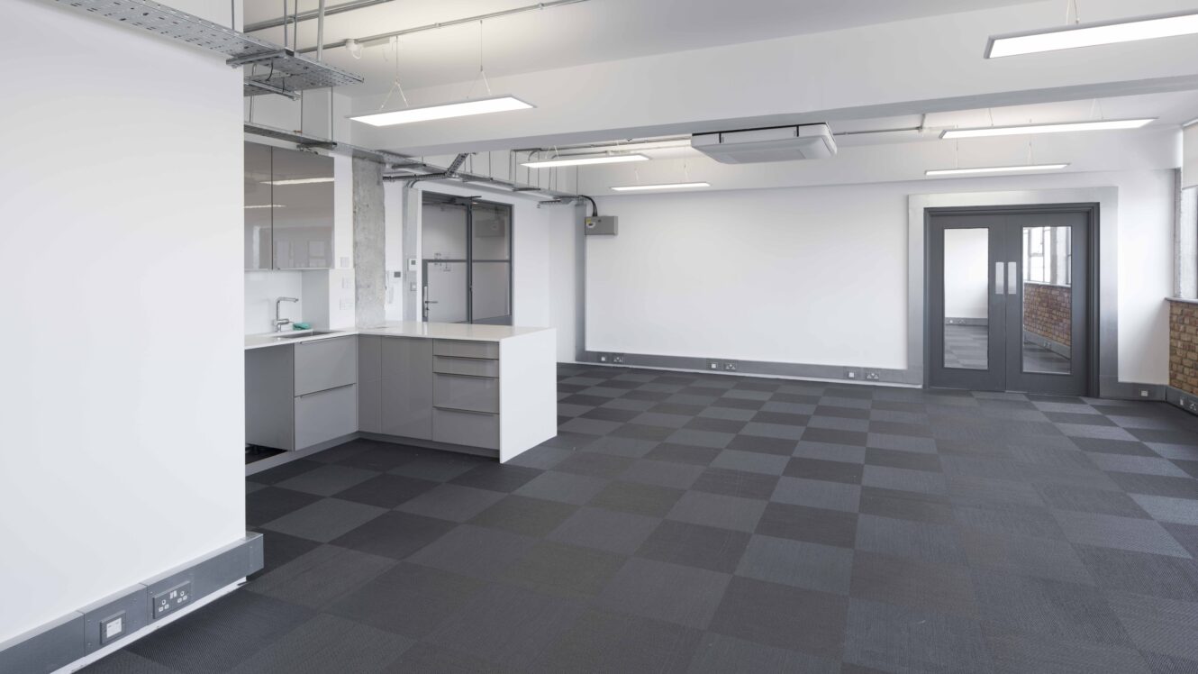 Modern empty office space with checkered gray carpet, a small kitchenette area, large windows, and white walls. Ceiling features exposed ducts and fluorescent lighting.