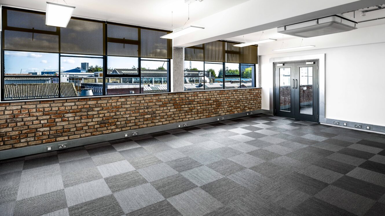 An empty office with carpeted flooring, brick accent wall, large windows, and double glass doors, illuminated by ceiling lights.