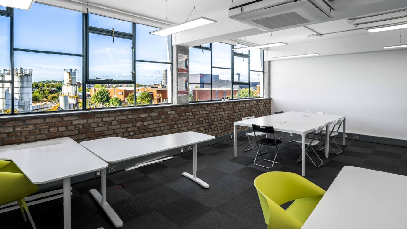 A modern classroom with white desks, black and yellow chairs, large windows, brick wall, and a view of industrial buildings and trees outside.
