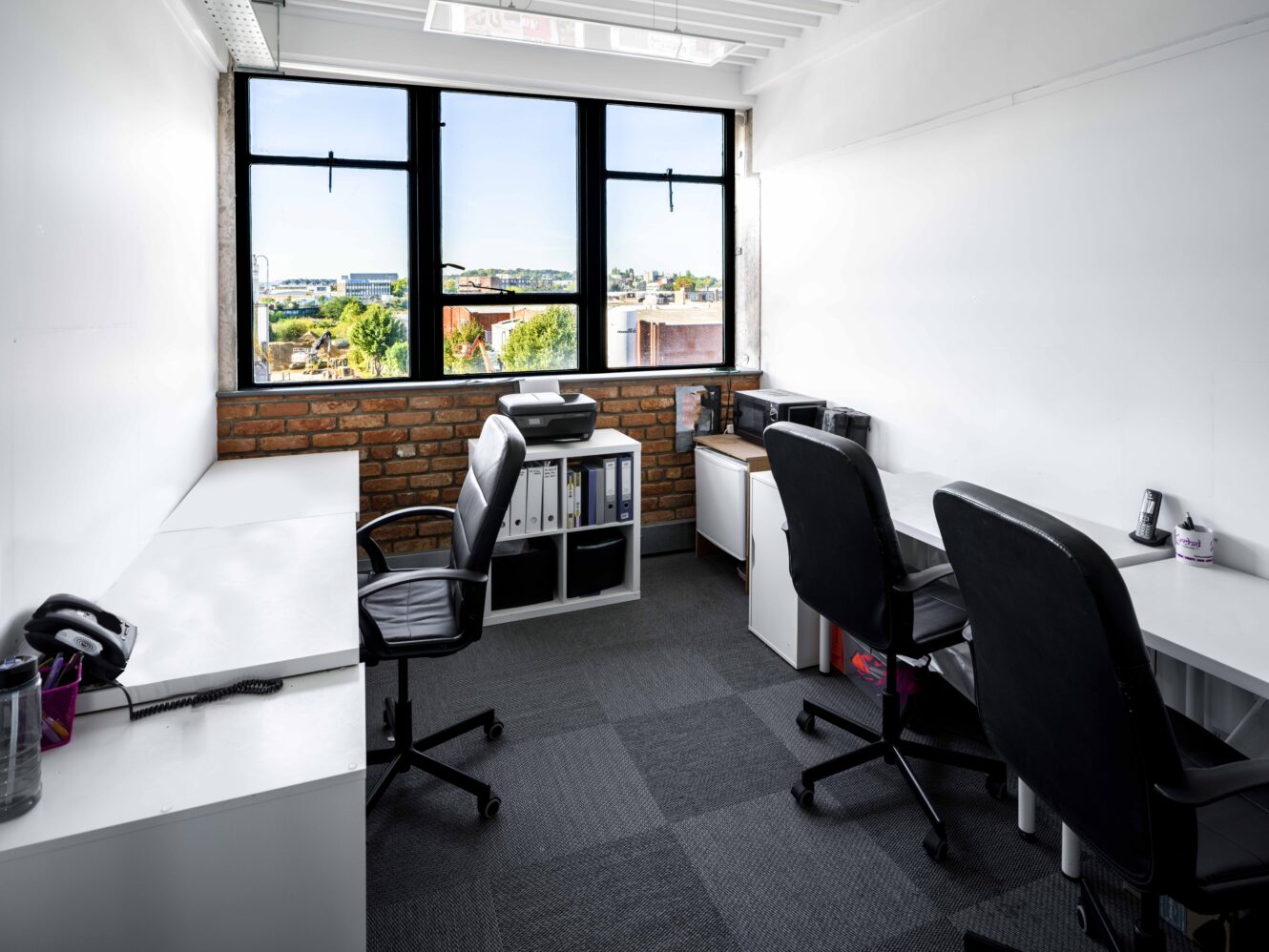 Small modern office with three desks, black chairs, a printer, office supplies, and large windows overlooking a cityscape.