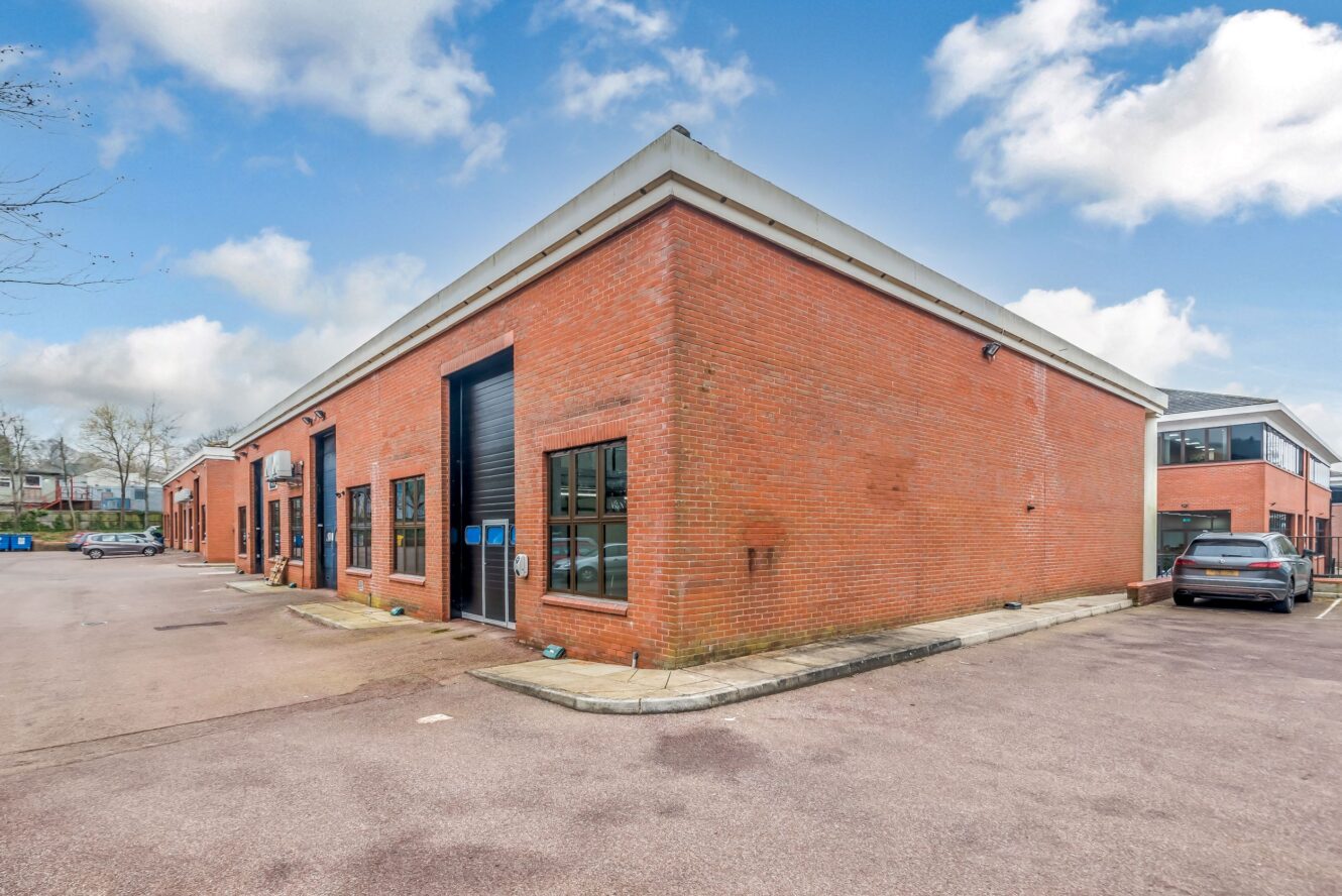 A modern red brick commercial building with large windows and a black garage door, situated in a paved parking area under a partly cloudy sky.