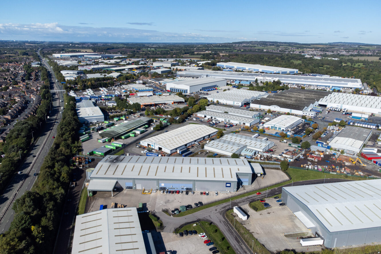 Aerial view of an industrial area with multiple warehouses, storage yards, parked trucks, and nearby roads, surrounded by fields and residential neighborhoods.