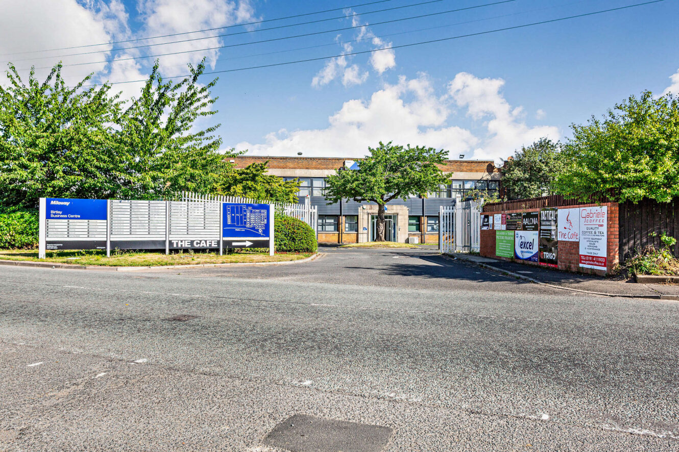 Entrance to an industrial estate with a gated driveway, several business signs, and a two-story brick building in the background.