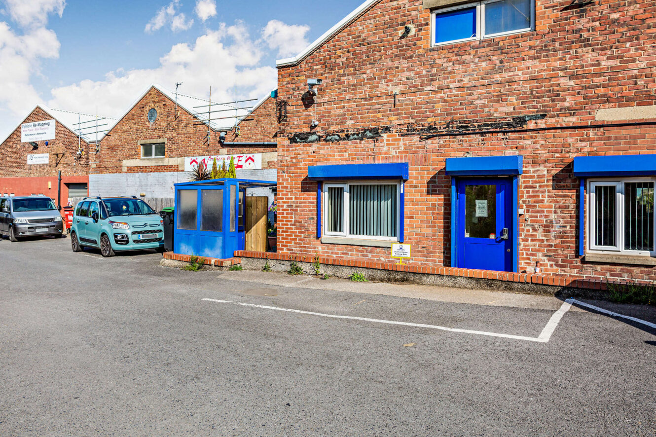 A brick industrial building with blue-framed windows and doors, a small blue booth, and several parked cars on an adjacent street under a partly cloudy sky.