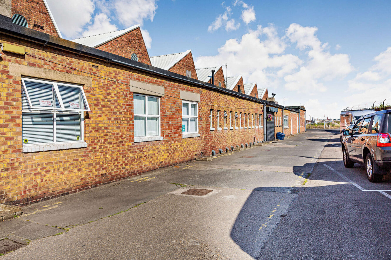 A row of single-story brick industrial buildings with white-framed windows, seen from a paved parking lot on a sunny day.