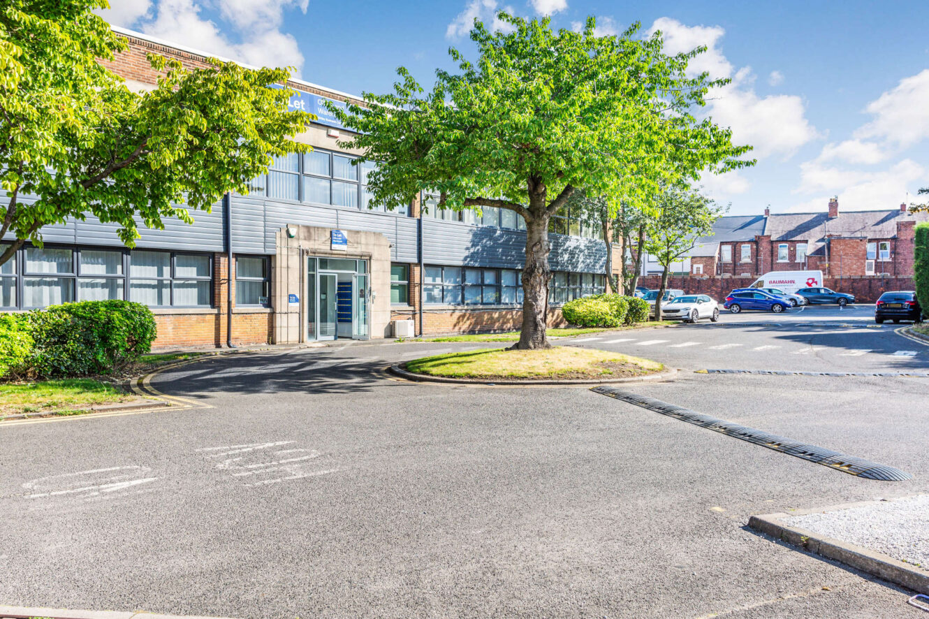 A commercial building with a blue sign, large windows, and a glass door is surrounded by trees, bushes, and parked cars on a sunny day.