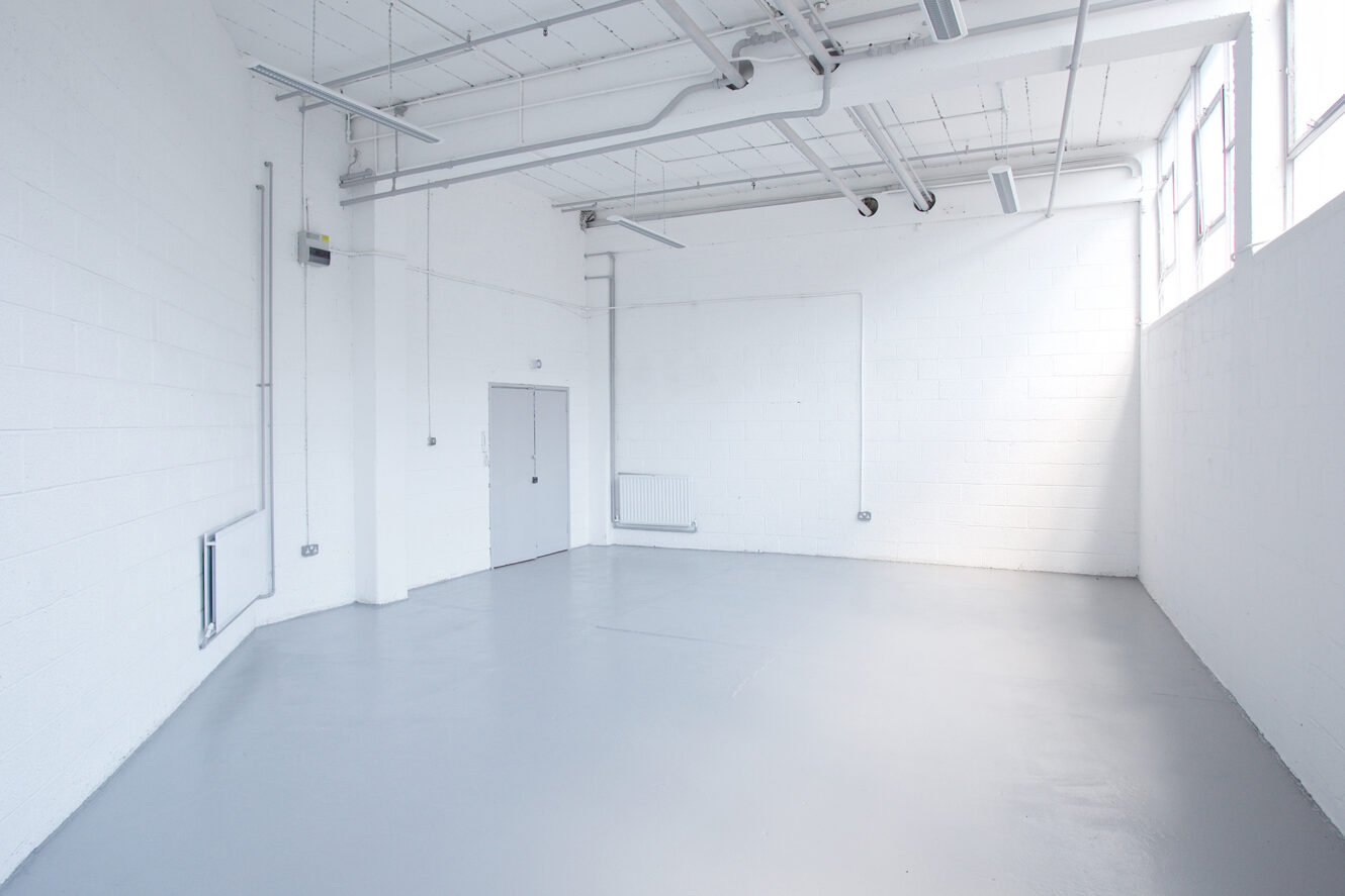 Empty industrial room with white brick walls, grey double doors, large windows, exposed pipes, and a smooth grey floor. Bright natural light fills the space.
