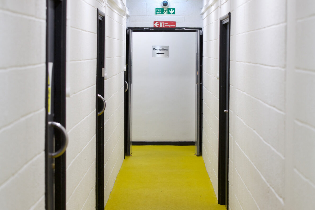 A hallway with white walls and yellow carpet leads to an open door labeled Reception with an arrow, below an emergency exit sign and fire extinguisher symbol.