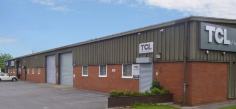 A row of industrial warehouse units with brown brick and metal siding; the front unit has TCL signage and a roller shutter door. Cars are parked in front of the building.