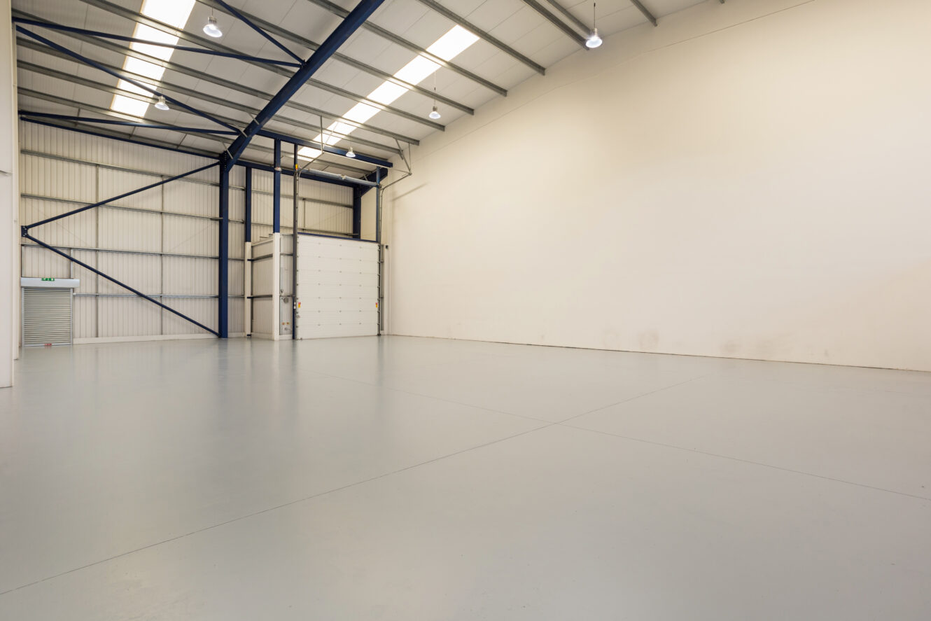 Interior of an empty, clean industrial warehouse with high ceilings, white walls, large overhead doors, and exposed metal beams.