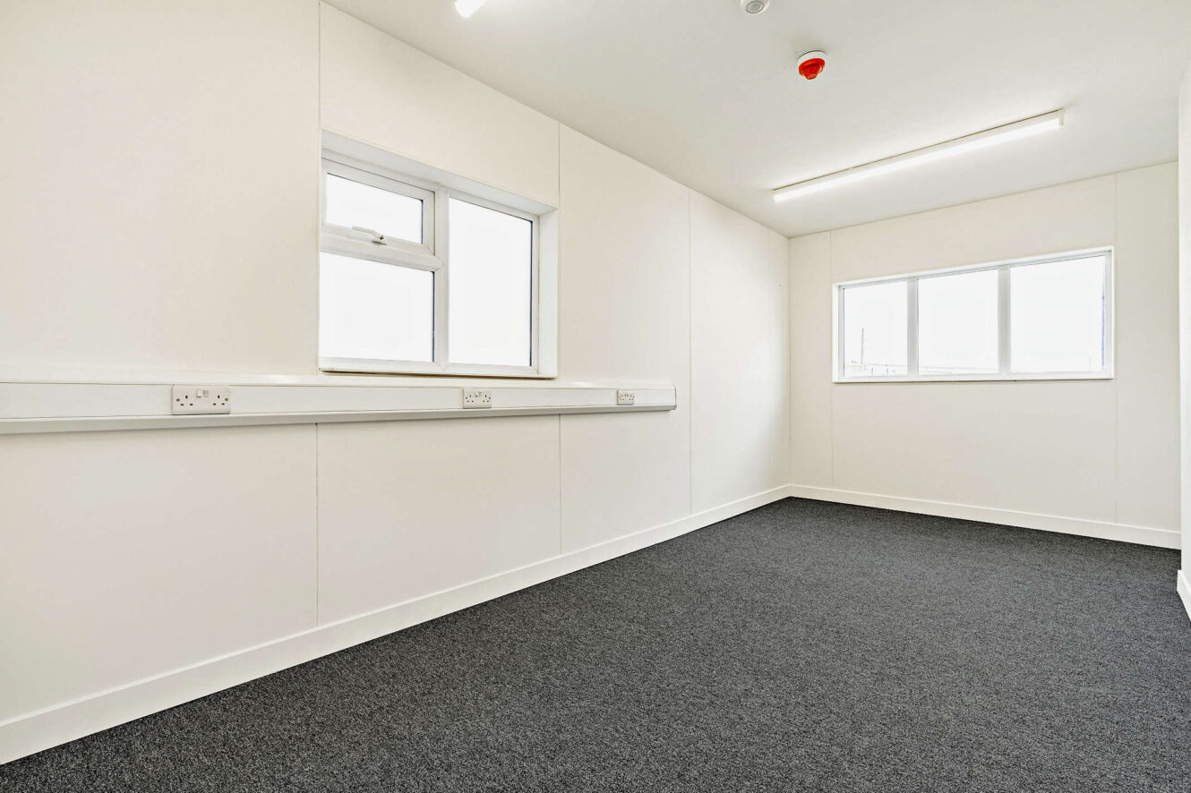 Empty office room with white walls, dark carpet, two windows, electrical outlets along one wall, and fluorescent ceiling lighting.