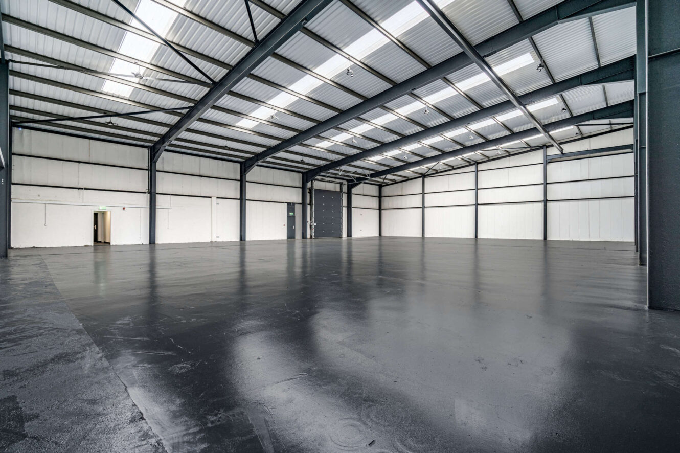 Interior of a large empty warehouse with polished dark floor, high metal ceiling, exposed beams, and white paneled walls. Light enters through windows and open doors.