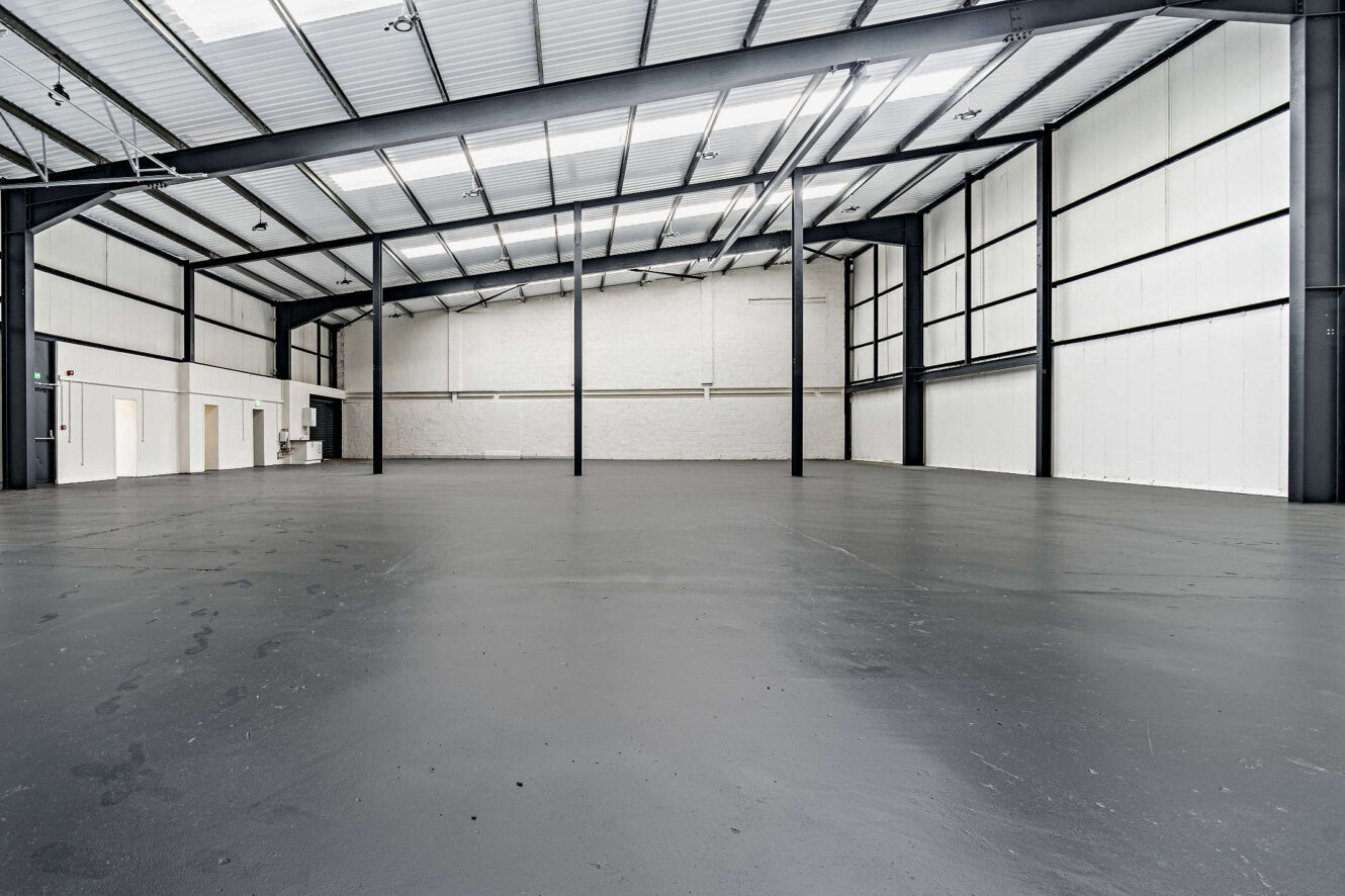 Interior of an empty industrial warehouse with high ceilings, metal beams, white walls, large open floor space, and closed doors visible on the left side.