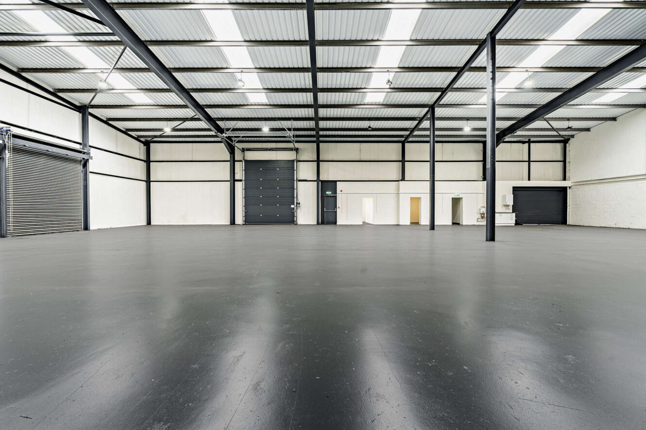 Spacious empty warehouse with black floor, white walls, exposed beams, large roller doors, and overhead lighting.