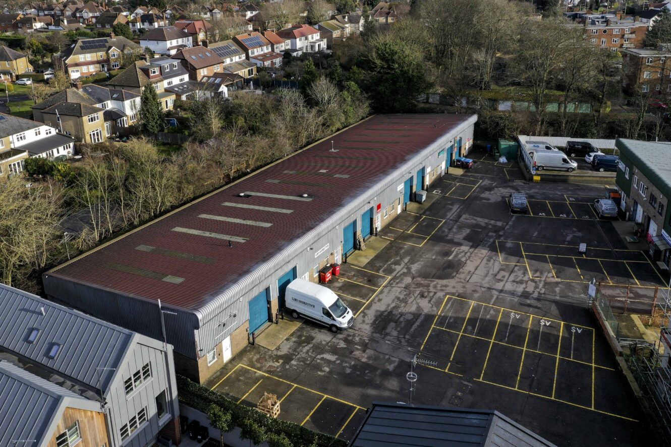 Aerial view of a warehouse building with blue doors, a white van parked outside, and empty parking spaces, surrounded by houses and trees.