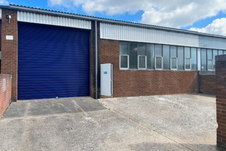 A brick industrial building with a large blue roller shutter door, several windows, and a concrete driveway under a partly cloudy sky.