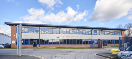 A modern, two-story office building with large windows, brick and panel exterior, located next to a road with parked cars and a blue fence under a partly cloudy sky.
