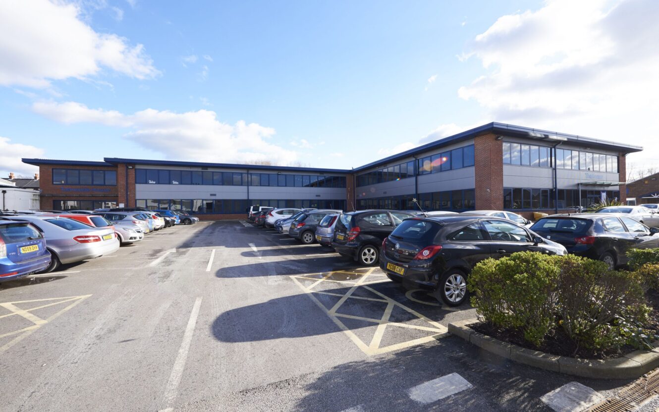 A modern two-story office building with large windows, surrounded by a parking lot filled with parked cars under a partly cloudy sky.