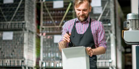 A man wearing an apron places a wine bottle into a box in a warehouse with metal racks in the background.