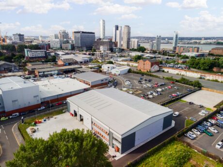 A large industrial warehouse with a parking lot is surrounded by other commercial buildings, with a city skyline visible in the background under a partly cloudy sky.