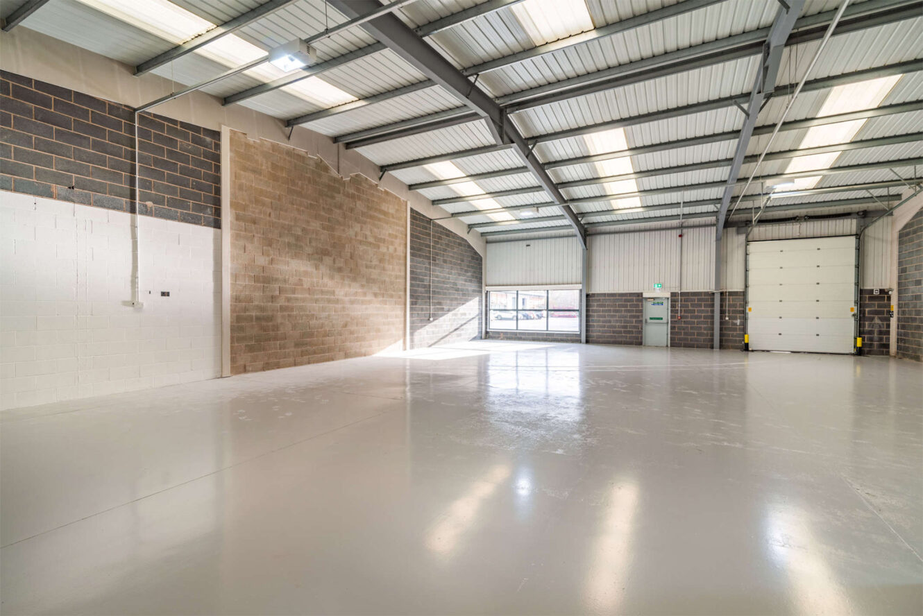 Spacious empty industrial warehouse with concrete floor, brick walls, high ceiling, large windows, and a roller shutter door.