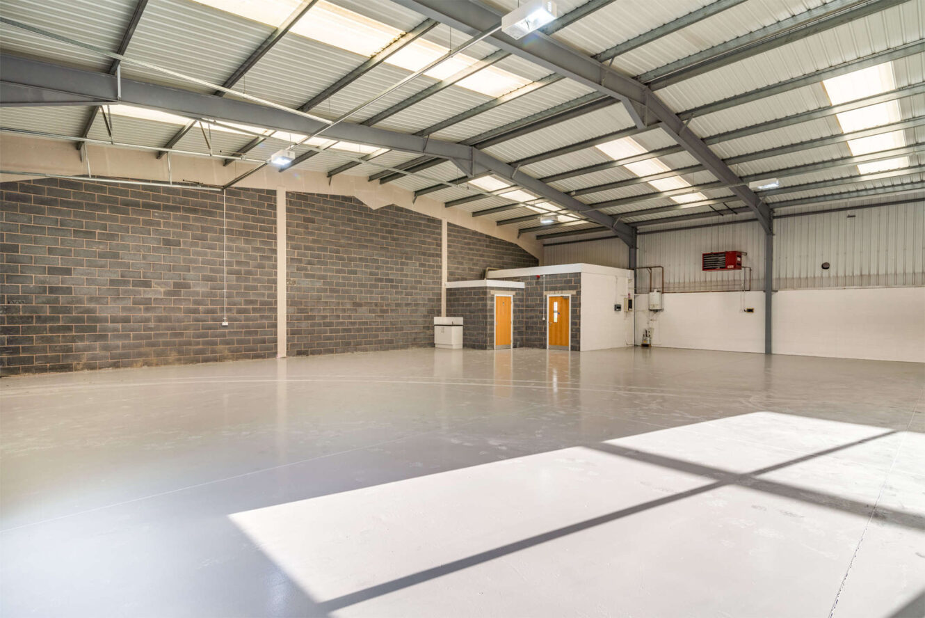Empty industrial warehouse with polished concrete floor, high ceiling, brick and metal walls, fluorescent lighting, and small office area with two wooden doors.