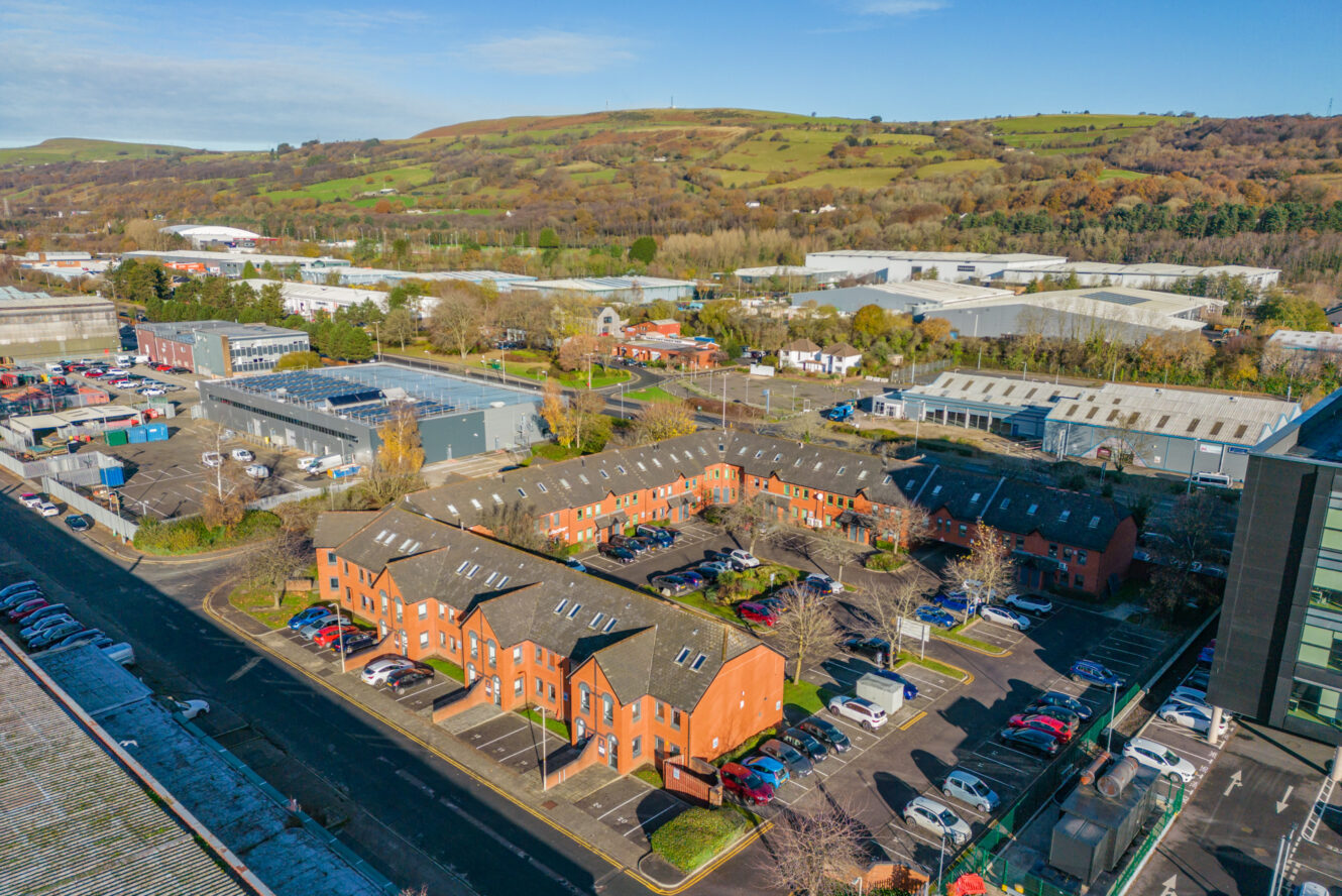 Aerial view of a business park with red-brick office buildings, parking lots, and surrounding greenery under a clear blue sky.