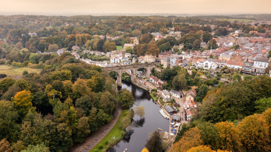 Aerial view of a stone bridge crossing a river, surrounded by trees with autumn foliage and a town with clustered buildings in the background.
