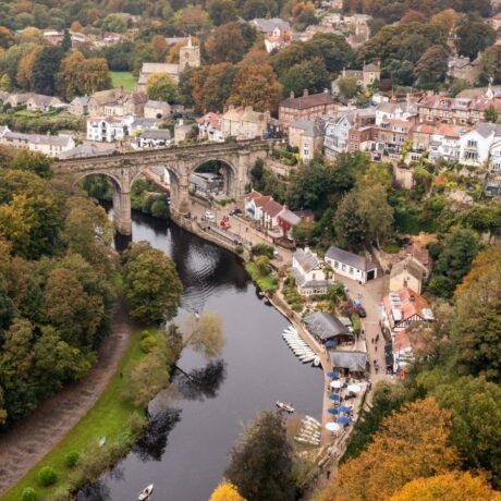 Aerial view of a historic stone bridge over a river, surrounded by trees and buildings in a small town. Boats are docked along the riverbank.