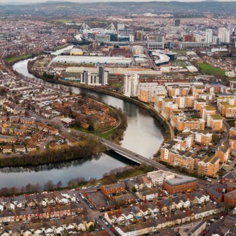 Aerial view of a cityscape with a river winding through residential and commercial buildings, a stadium in the background, and roads connecting various neighborhoods.