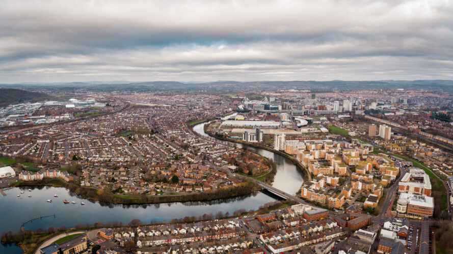 Aerial view of a city with a river running through it, surrounded by residential buildings, roads, and overcast skies.