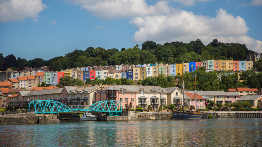 Colorful houses on a hillside overlook a waterfront with a blue bridge, moored boats, and trees under a partly cloudy sky.