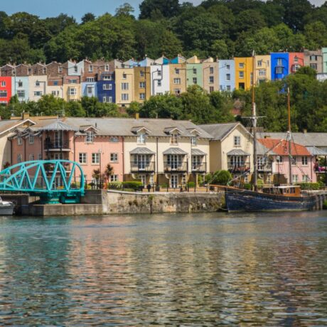 Colorful row houses line a hillside above the harbor, with a turquoise footbridge and docked boats reflected in the water below.