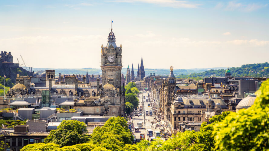 A cityscape view of Edinburgh, Scotland, featuring the Balmoral Hotel clock tower, historic buildings, and greenery under a clear sky.