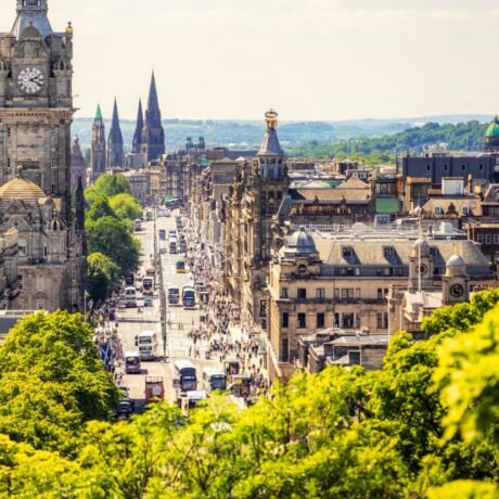 A busy city street lined with historic buildings, buses, and pedestrians, with church spires and greenery visible under a clear sky.