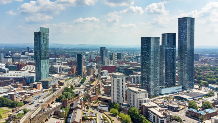 Aerial view of a cityscape with modern skyscrapers, mid-rise buildings, roads, and rail tracks under a partly cloudy sky.