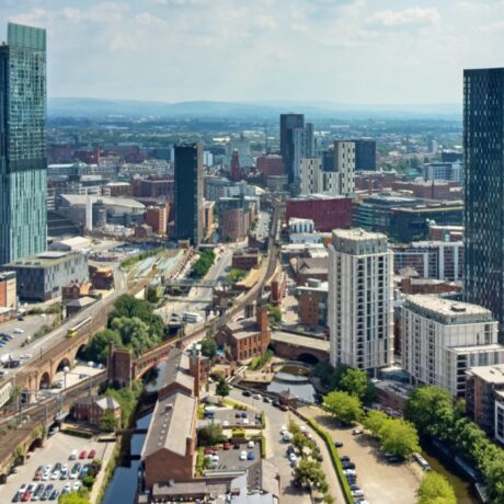 Aerial view of a modern cityscape with tall buildings, railway lines, roads, and a canal on a partly cloudy day.