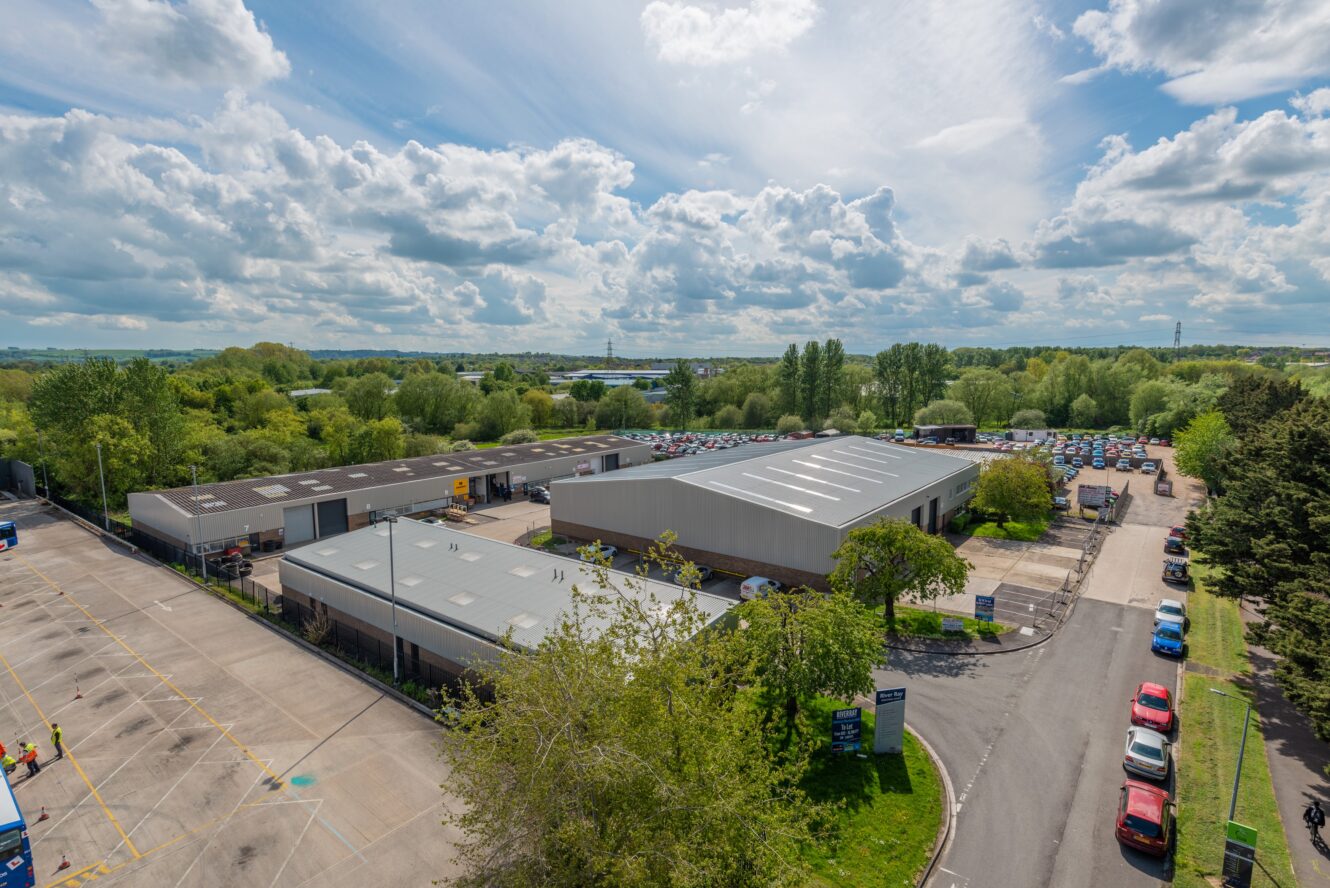 Aerial view of an industrial complex with several warehouse buildings, a parking lot, and surrounding greenery under a partly cloudy sky.