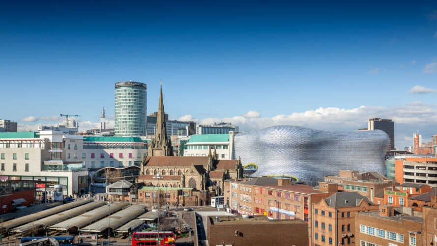 Cityscape of Birmingham, UK, featuring the Bullring shopping centre with its metallic facade, historic church, modern buildings, and a busy street with buses and market stalls.