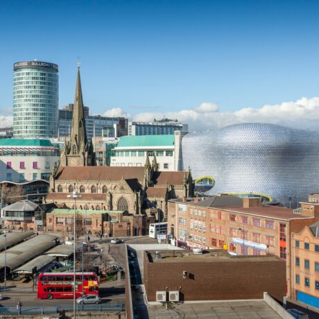 Cityscape view of Birmingham, UK, featuring a church, modern buildings including the Bullring shopping centre, a double-decker bus, and clear blue sky.