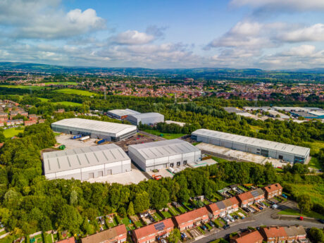 Aerial view of a group of large industrial warehouses surrounded by trees, with residential houses in the foreground and a town in the background.