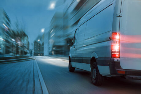 A white delivery van drives quickly down a city street at dusk, with its brake lights on and buildings blurred in the background.