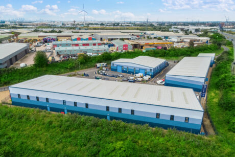 Aerial view of an industrial estate with several large warehouses, office units, parked vehicles, and surrounding greenery under a partly cloudy sky.
