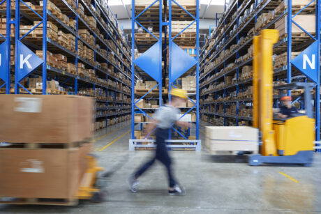 Warehouse interior with tall shelves of boxes, two workers in motion, and a yellow forklift moving a pallet in the aisle.