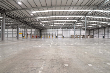 Interior of a large, empty warehouse with high ceilings, exposed beams, concrete floor, and rows of loading bay doors along the far wall.