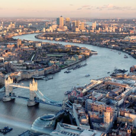 Aerial view of London showing Tower Bridge over the River Thames, with city buildings and Canary Wharf in the background.