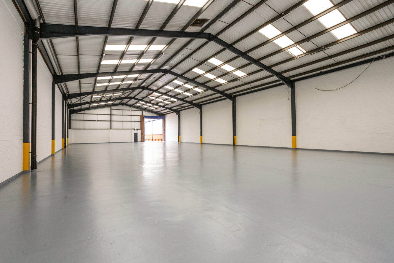 Interior of a large, empty warehouse with white walls, high ceiling, skylights, and a gray concrete floor.