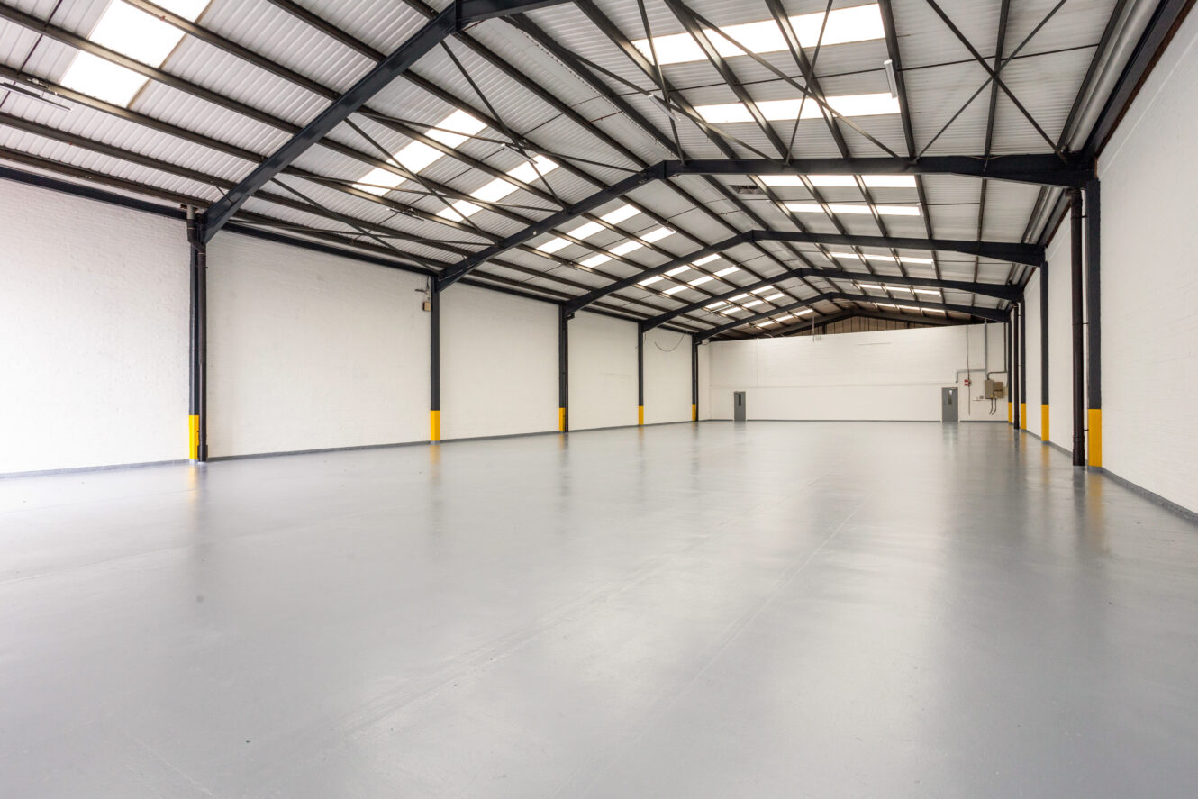 Large, empty industrial warehouse with white walls, gray floor, high ceiling, exposed steel beams, and skylights letting in natural light.