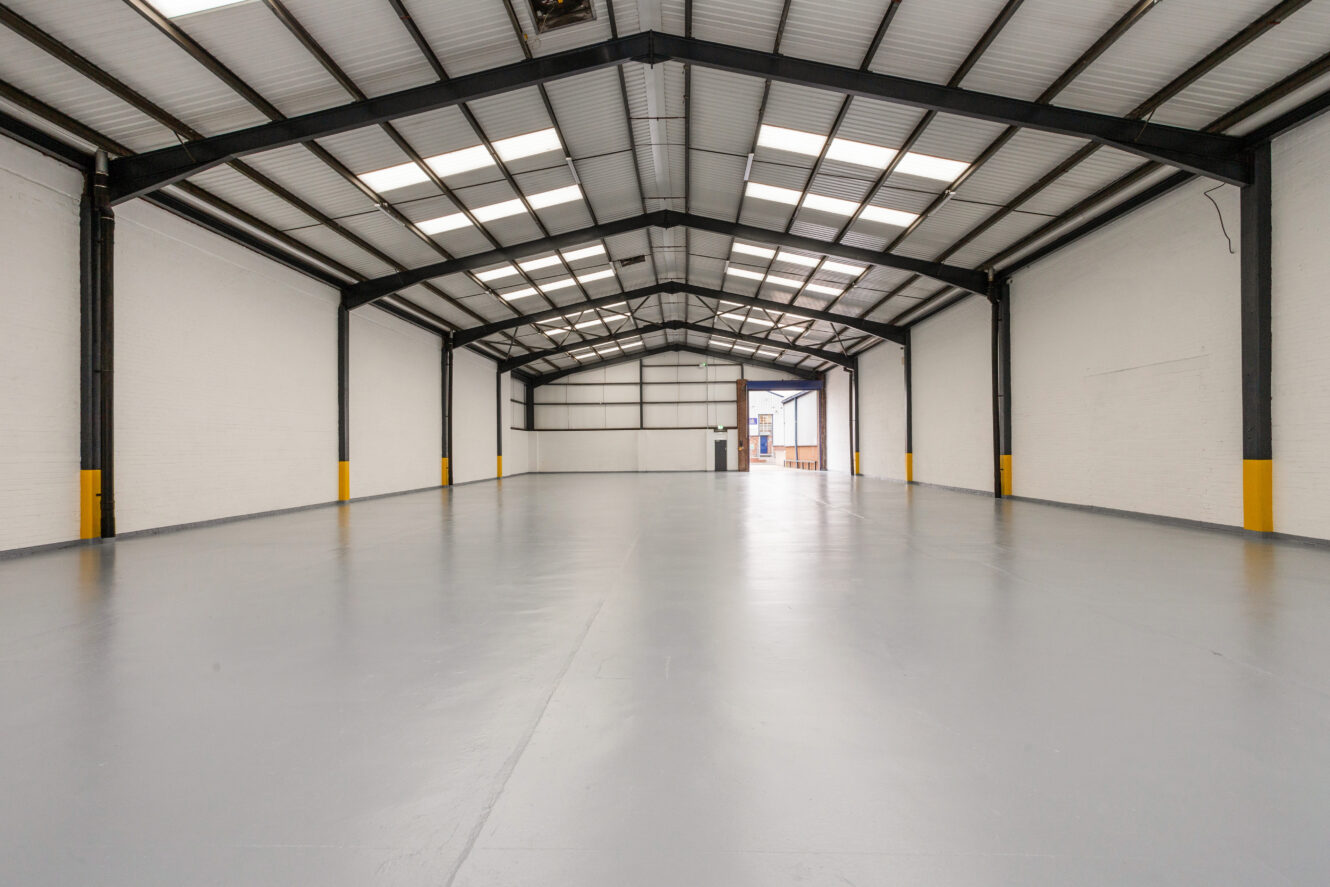 Wide, empty industrial warehouse with white walls, high ceiling, exposed metal beams, and polished gray concrete floor. Yellow markers are visible on the lower wall columns.